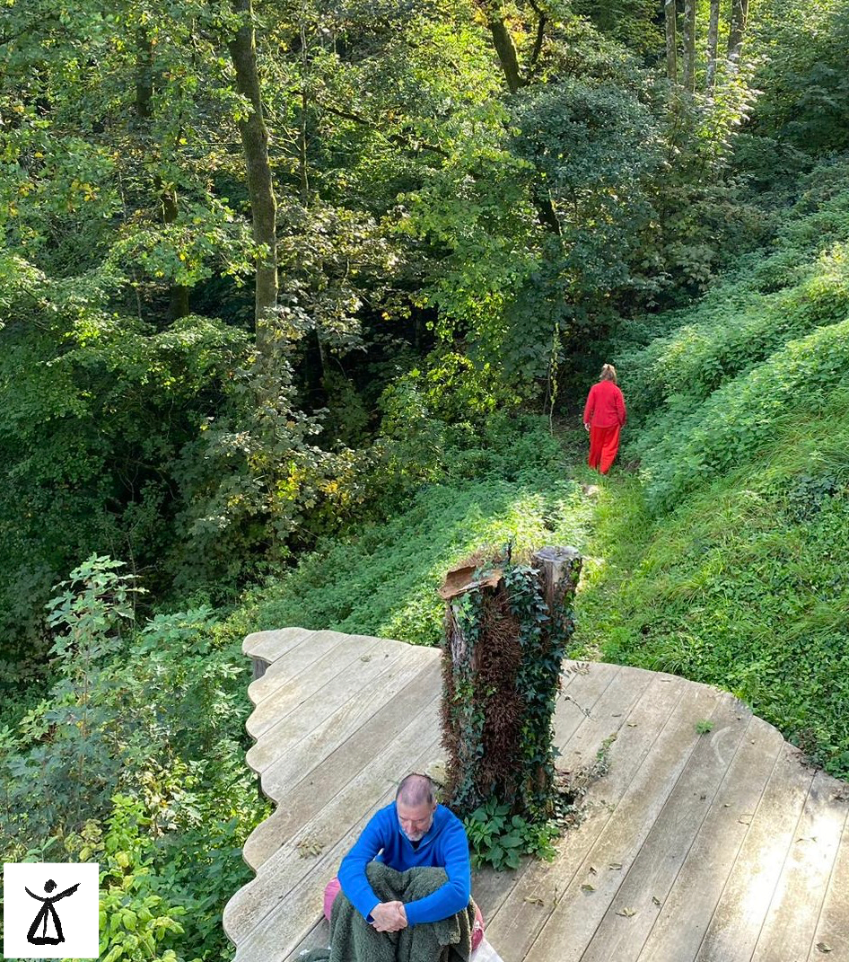 Un homme assis habillé en bleu; dans son dos, une femme en rouge s'éloigne dans la forêt